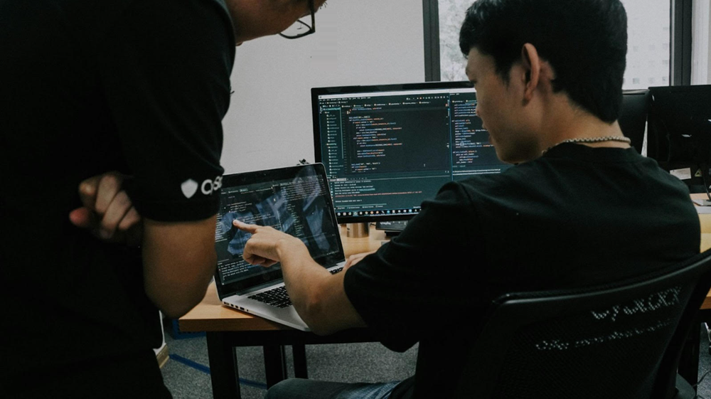An image of a man sitting at a desk with a laptop and external screen